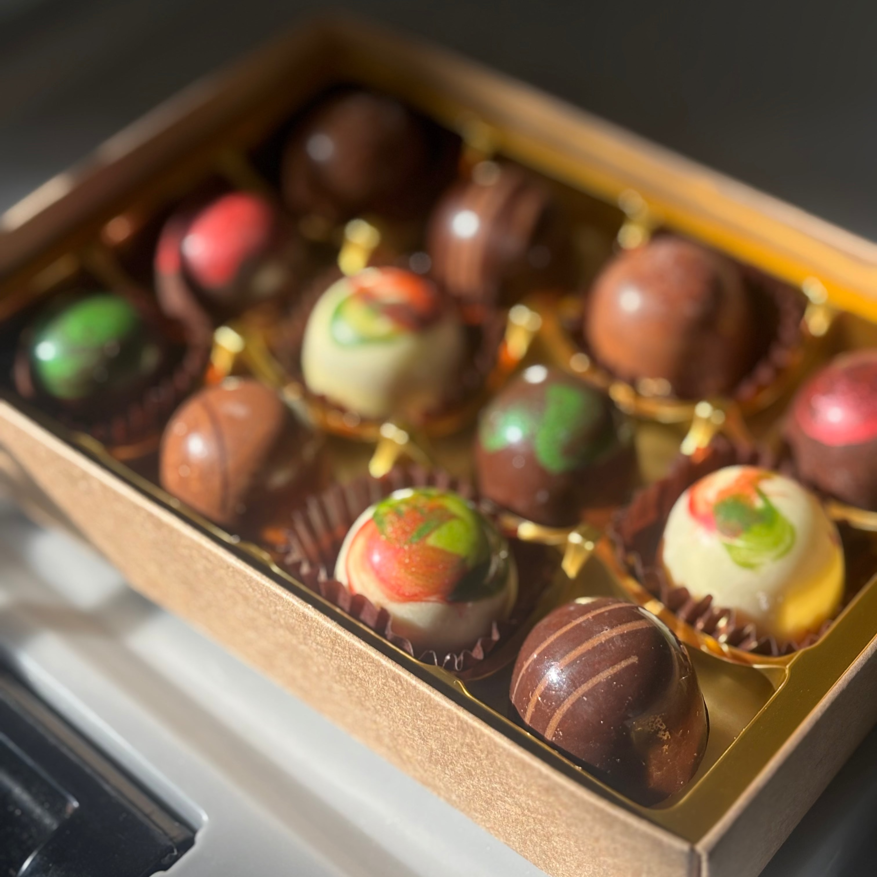 An open box of assorted gourmet chocolates with gold foil detailing, featuring various chocolate truffles and milk chocolate pieces in brown, white, and red colors.